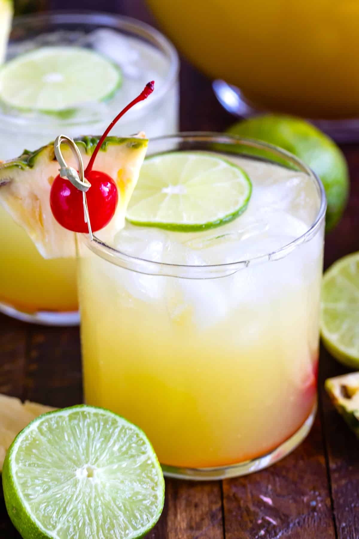 yellow drink in a short clear glass with cherries and pineapple slices and limes on the rim.