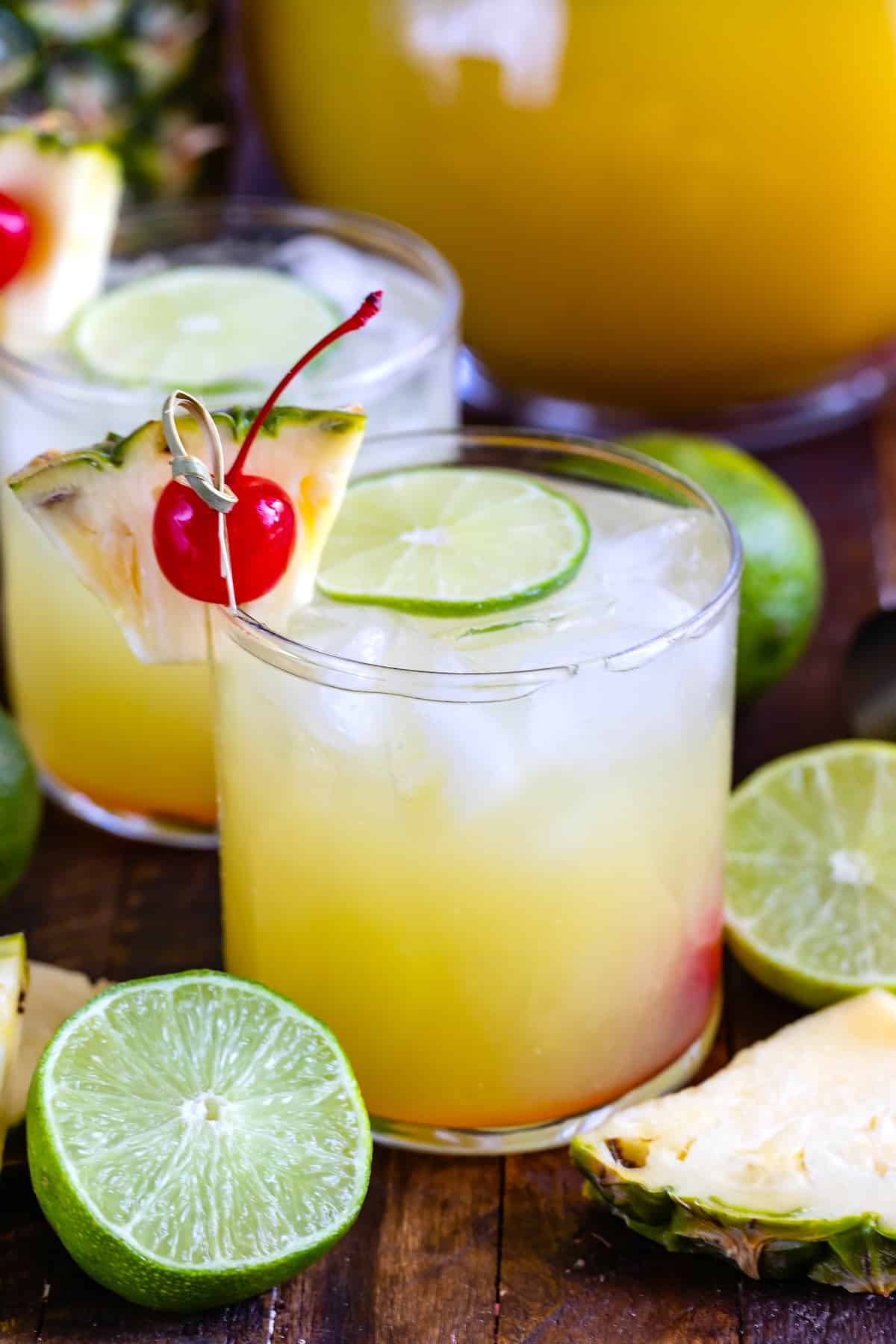 yellow drink in a short clear glass with cherries and pineapple slices and limes on the rim.