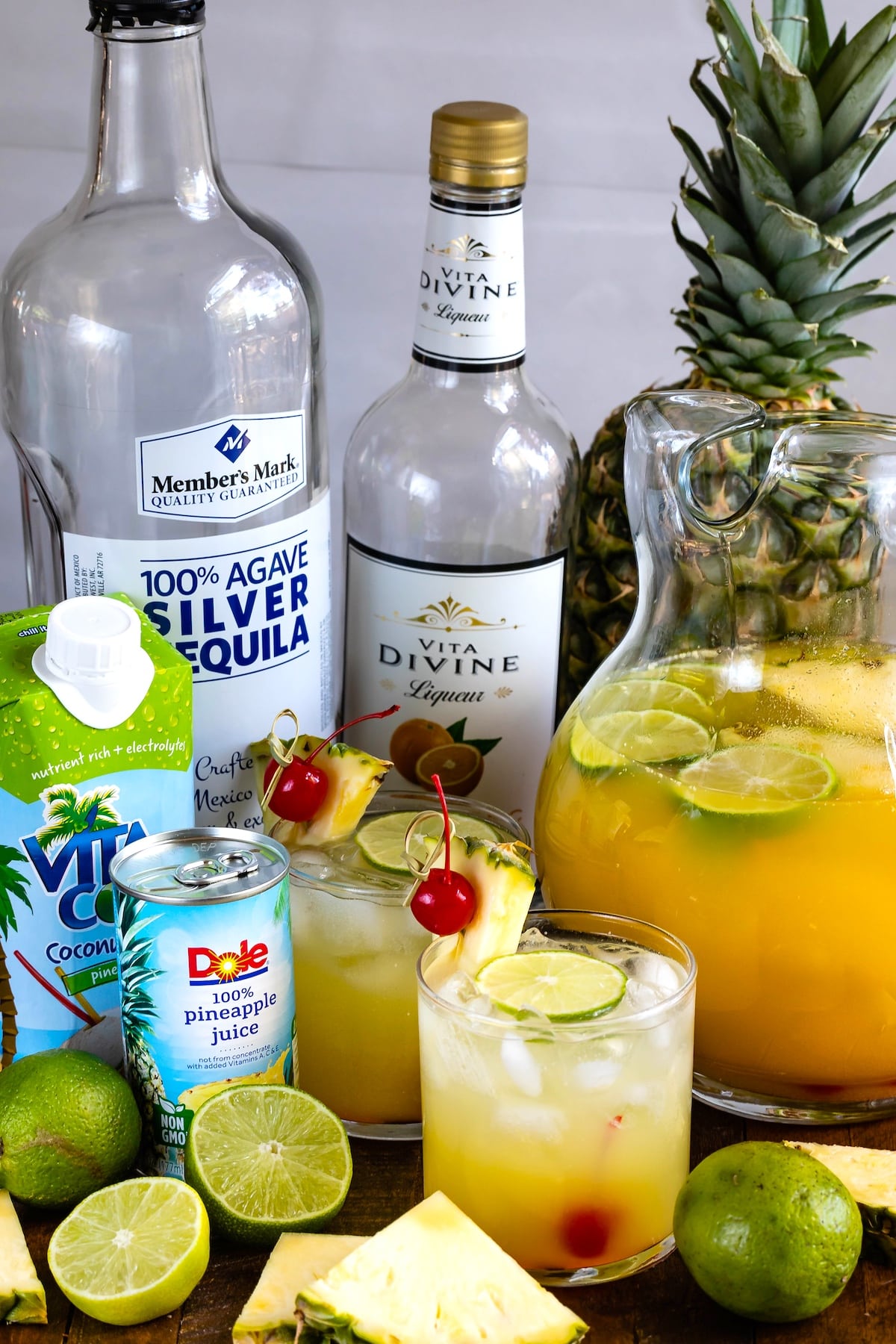 yellow drink in a clear glass with cherries and pineapple slices and limes on the rim and with the ingredients behind them.