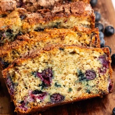 Close-up of sliced Blueberry Banana Bread on a wooden cutting board. The bread features a golden-brown crust with cinnamon sugar on top and visible blueberries in every slice, with fresh blueberries scattered nearby.