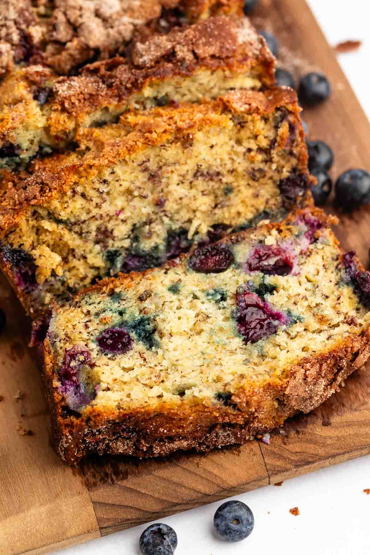 Close-up of Blueberry Banana Bread on a wooden board. The moist bread features visible blueberries and a golden-brown, slightly crumbly crust, with a few loose blueberries scattered nearby.