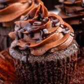 A close-up of a chocolate cupcake with rich chocolate frosting, topped with chocolate syrup. The cupcake wrapper is partially peeled back, revealing the moist, dark cake—a delicious glimpse into a perfect chocolate cupcake recipe. Other cupcakes blur in the background.