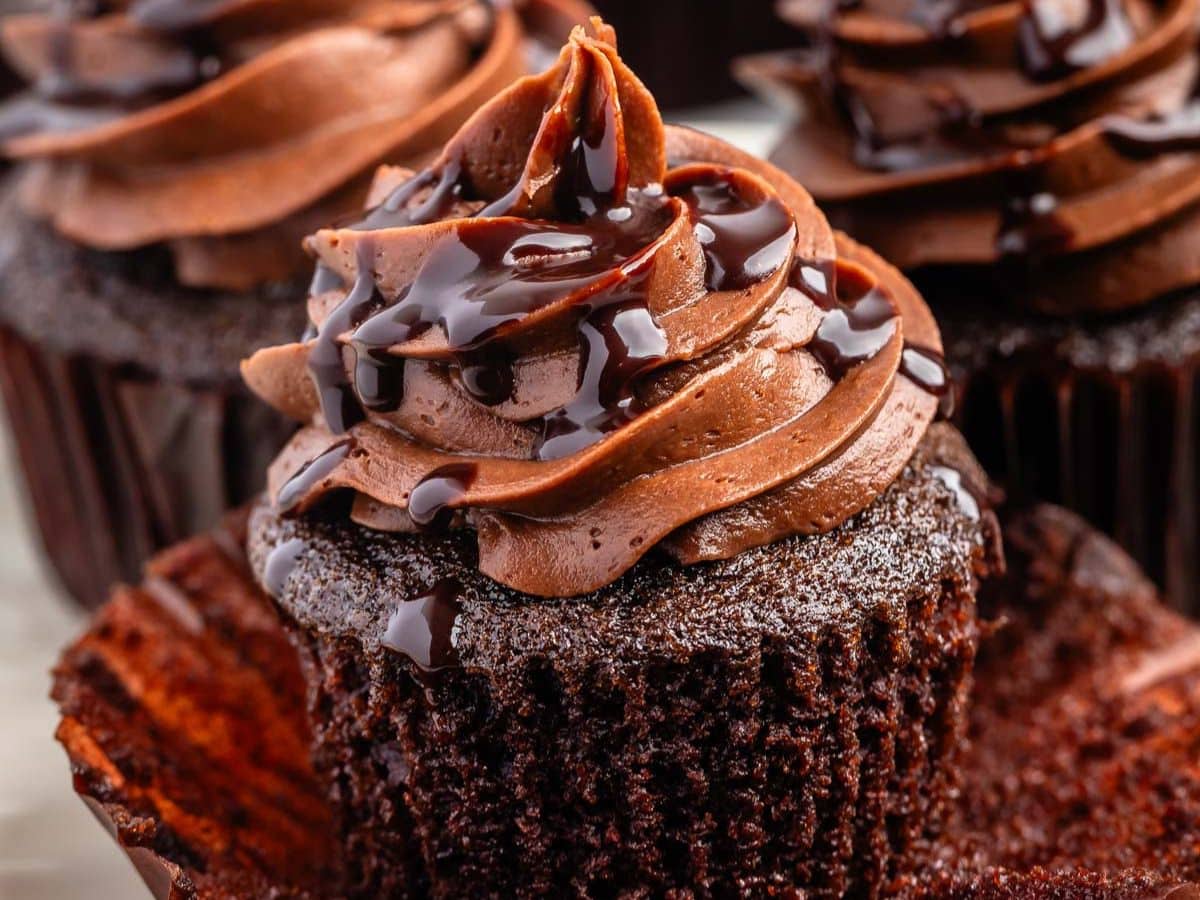 A close-up of a chocolate cupcake with rich chocolate frosting, topped with chocolate syrup. The cupcake wrapper is partially peeled back, revealing the moist, dark cake—a delicious glimpse into a perfect chocolate cupcake recipe. Other cupcakes blur in the background.