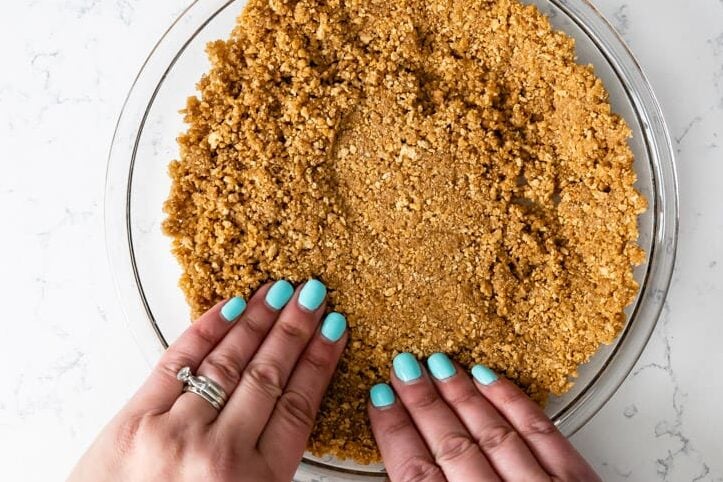 glass pie plate with graham cracker crust crumbs being pressed by hands.