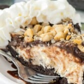 A slice of Hula Pie with a chocolate crust, topped with chopped nuts, chocolate sauce, and whipped cream sits on a white plate with a fork.