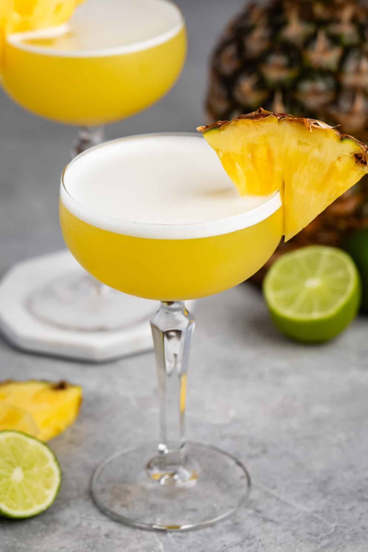 tall clear martini glass holding a yellow drink with a pineapple slice on the rim.