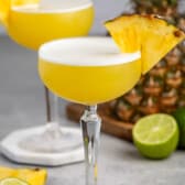 tall clear martini glass holding a yellow drink with a pineapple slice on the rim.