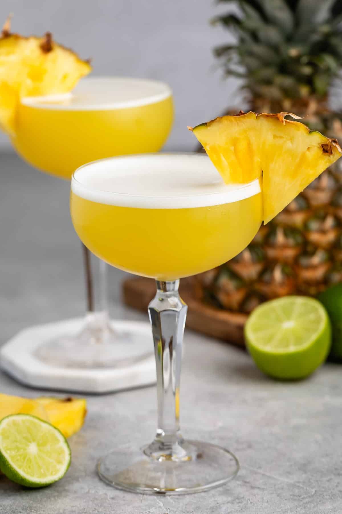 tall clear martini glass holding a yellow drink with a pineapple slice on the rim.