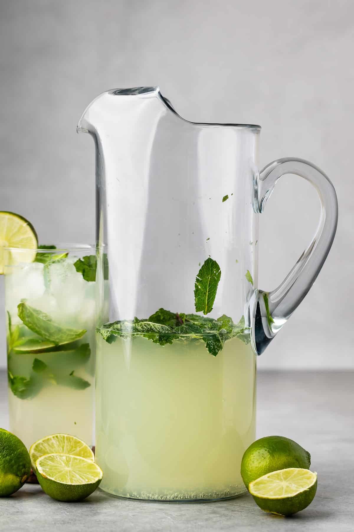 mojito in a tall clear pitcher with sliced limes in and around the glass with mint.