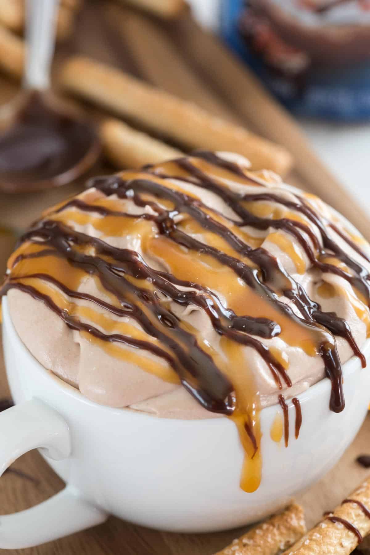 cheesecake dip in a white mug with caramel and chocolate drizzled over top.