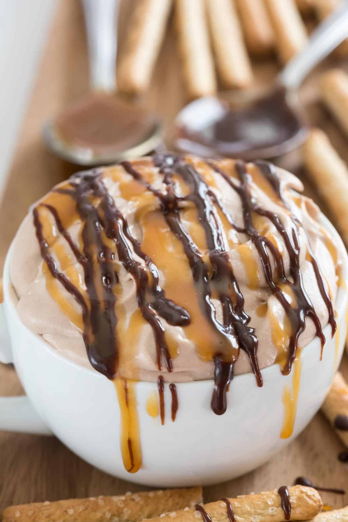 cheesecake dip in a white mug with caramel and chocolate drizzled over top.