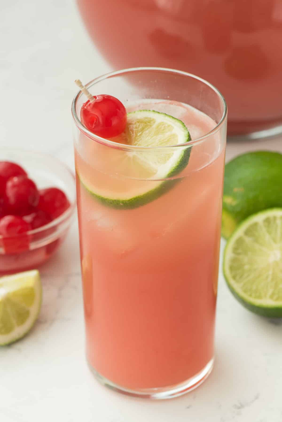pink drink in a tall clear glass with a lime and a cherry inside.
