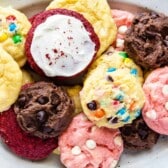 A plate filled with assorted cake mix cookies, including chocolate, red velvet with white frosting, yellow cake mix cookies with colorful candies, and pink cookies with white chocolate chips.