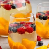 clear glasses holding the clear drink with raspberries, blueberries, and peaches sitting inside.