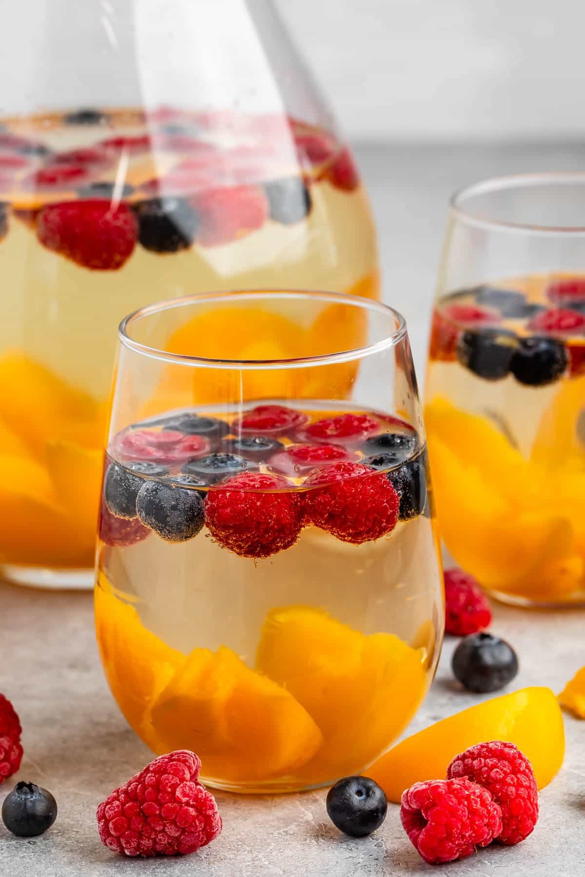 clear glasses holding the clear drink with raspberries, blueberries, and peaches sitting inside.
