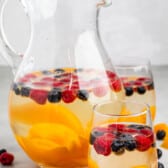 clear glasses holding the clear drink with raspberries, blueberries, and peaches sitting inside.