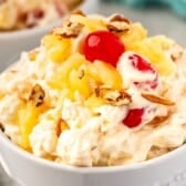 A close-up of a bowl filled with creamy pineapple fluff fruit salad, topped with pineapple chunks, maraschino cherries, and pecan pieces. The salad appears fluffy and colorful.