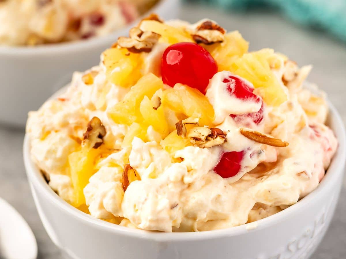 A close-up of a bowl filled with creamy pineapple fluff fruit salad, topped with pineapple chunks, maraschino cherries, and pecan pieces. The salad appears fluffy and colorful.