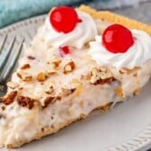 A slice of creamy millionaire pie with chopped nuts, topped with two swirls of whipped cream and maraschino cherries, served on a white plate with a fork.