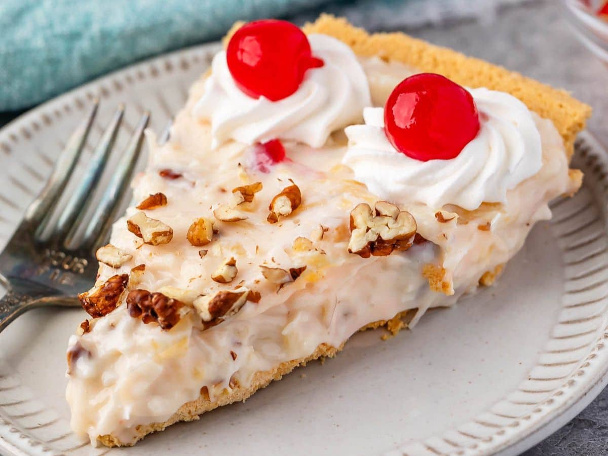 A slice of creamy millionaire pie with chopped nuts, topped with two swirls of whipped cream and maraschino cherries, served on a white plate with a fork.