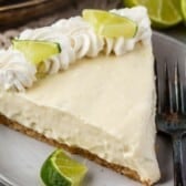A slice of no bake key lime pie with a graham cracker crust, topped with whipped cream and lime wedges, sits on a plate with a fork and extra lime slices on the side.
