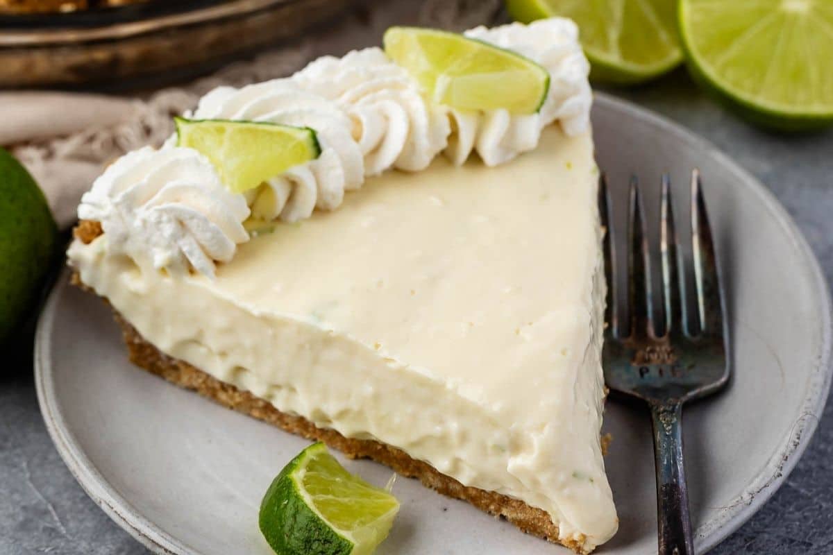 A slice of no bake key lime pie with a graham cracker crust, topped with whipped cream and lime wedges, sits on a plate with a fork and extra lime slices on the side.