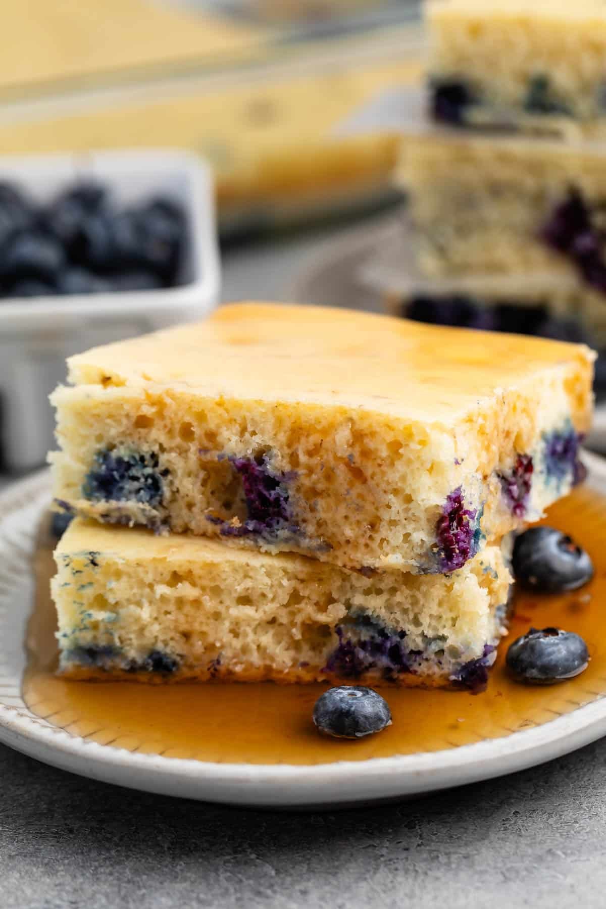 stacked square pancakes with blueberries baked in on a white plate covered in syrup.