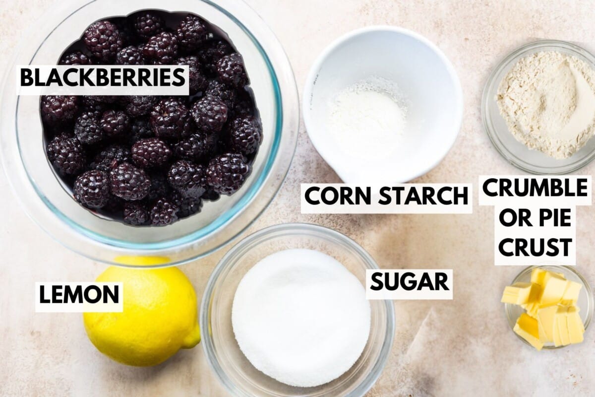 Top-down view of baking ingredients for Blackberry Pie labeled: a bowl of blackberries, a whole lemon, sugar, corn starch, crumble or pie crust, and butter pieces on a light surface.