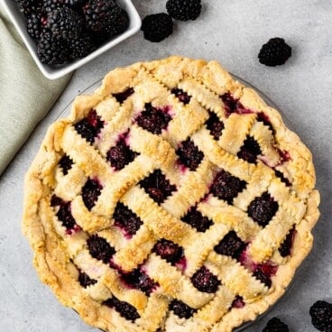 A freshly baked Blackberry Pie with a golden lattice crust sits on a gray surface, surrounded by loose blackberries and a small square dish filled with blackberries. A green cloth napkin is partially visible.