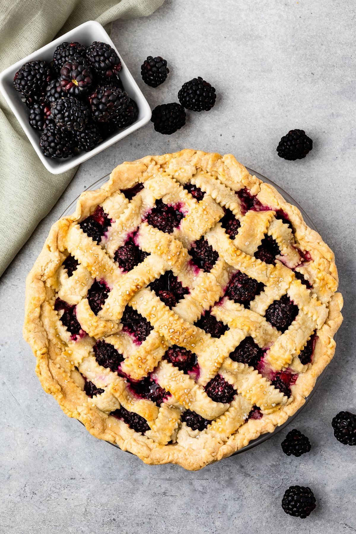 A freshly baked Blackberry Pie with a golden lattice crust sits on a gray surface, surrounded by loose blackberries and a small square dish filled with blackberries. A green cloth napkin is partially visible.