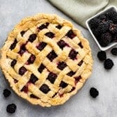 A freshly baked Blackberry Pie with a golden lattice crust sits on a gray surface. Several blackberries are scattered around, and a small white bowl filled with blackberries is nearby, next to a green cloth.