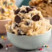 A bowl filled with chocolate chip edible cookie dough recipe, topped with extra chocolate chips. In the background, two more bowls hold different cookie doughs and colorful sprinkles are scattered on the table.