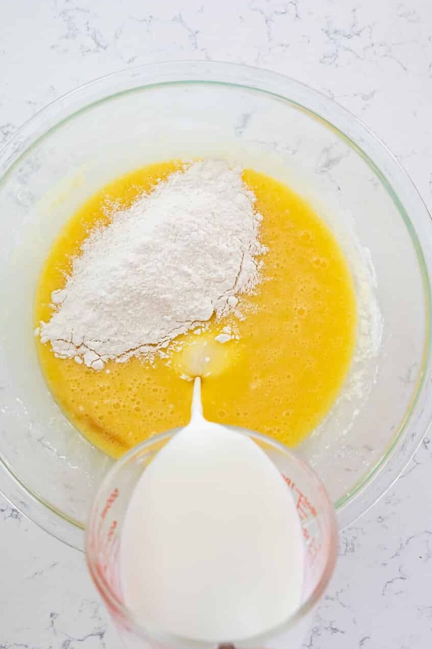 cake batter in glass bowl with flour and hand pouring milk into batter on marble background.