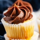 A close-up of a mini cupcake in a white paper liner, topped with a swirl of rich, creamy chocolate frosting. A blurred dark blue cloth is in the background.