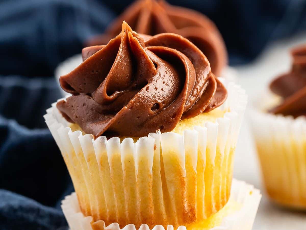 A close-up of a mini cupcake in a white paper liner, topped with a swirl of rich, creamy chocolate frosting. A blurred dark blue cloth is in the background.