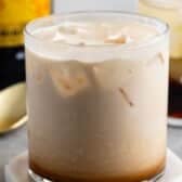 A glass of creamy iced cocktail, likely based on a classic White Russian recipe, sits on a coaster with visible ice cubes and layered colors. A gold spoon and blurred bottles are in the background.