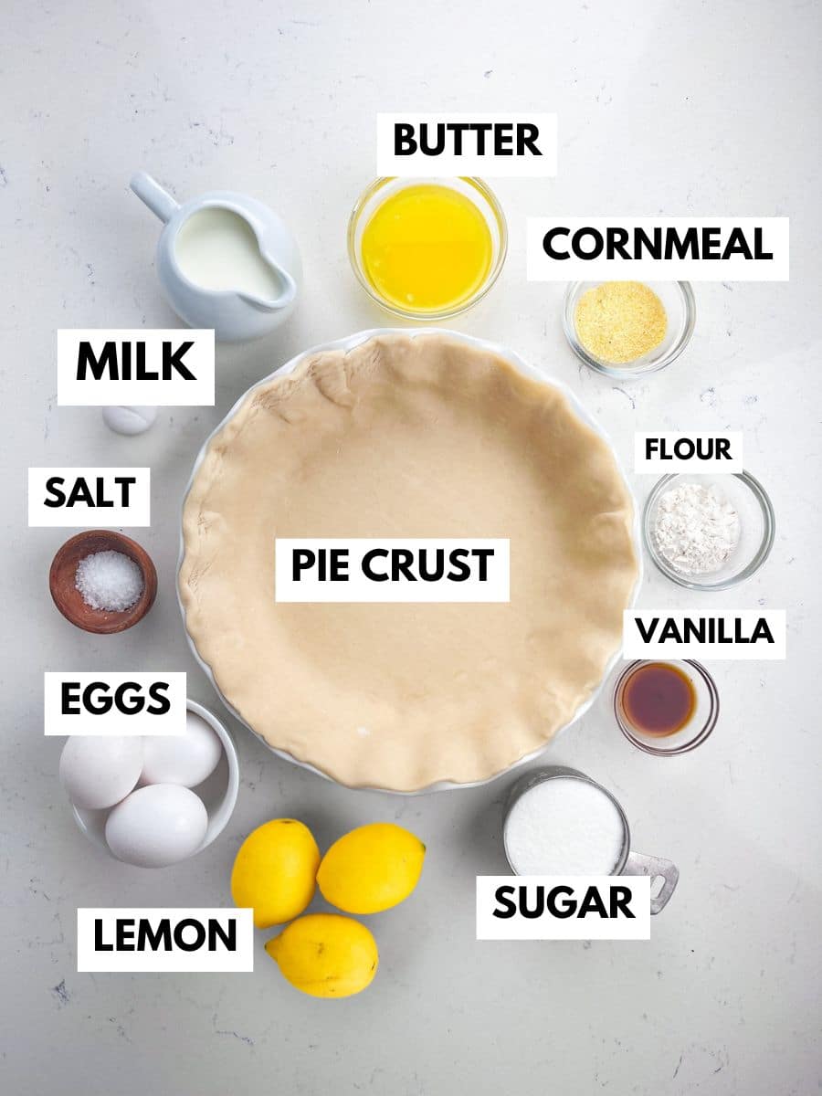 Overhead view of labeled baking ingredients for lemon chess pie: a pie crust in a dish surrounded by bowls of butter, cornmeal, sugar, flour, vanilla, eggs, salt, milk, and whole lemons on a white surface.