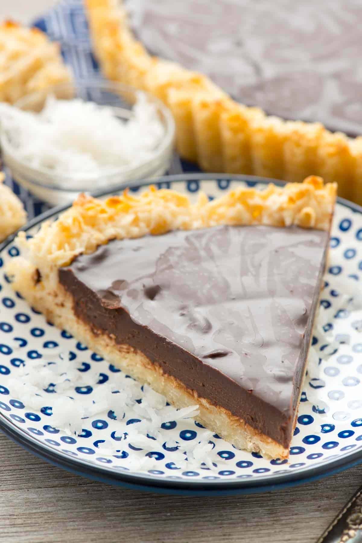 A slice of chocolate tart with a coconut crust sits on a white plate with blue dots. Shredded coconut is scattered around the plate. In the background, more tart slices and a bowl of shredded coconut are visible.