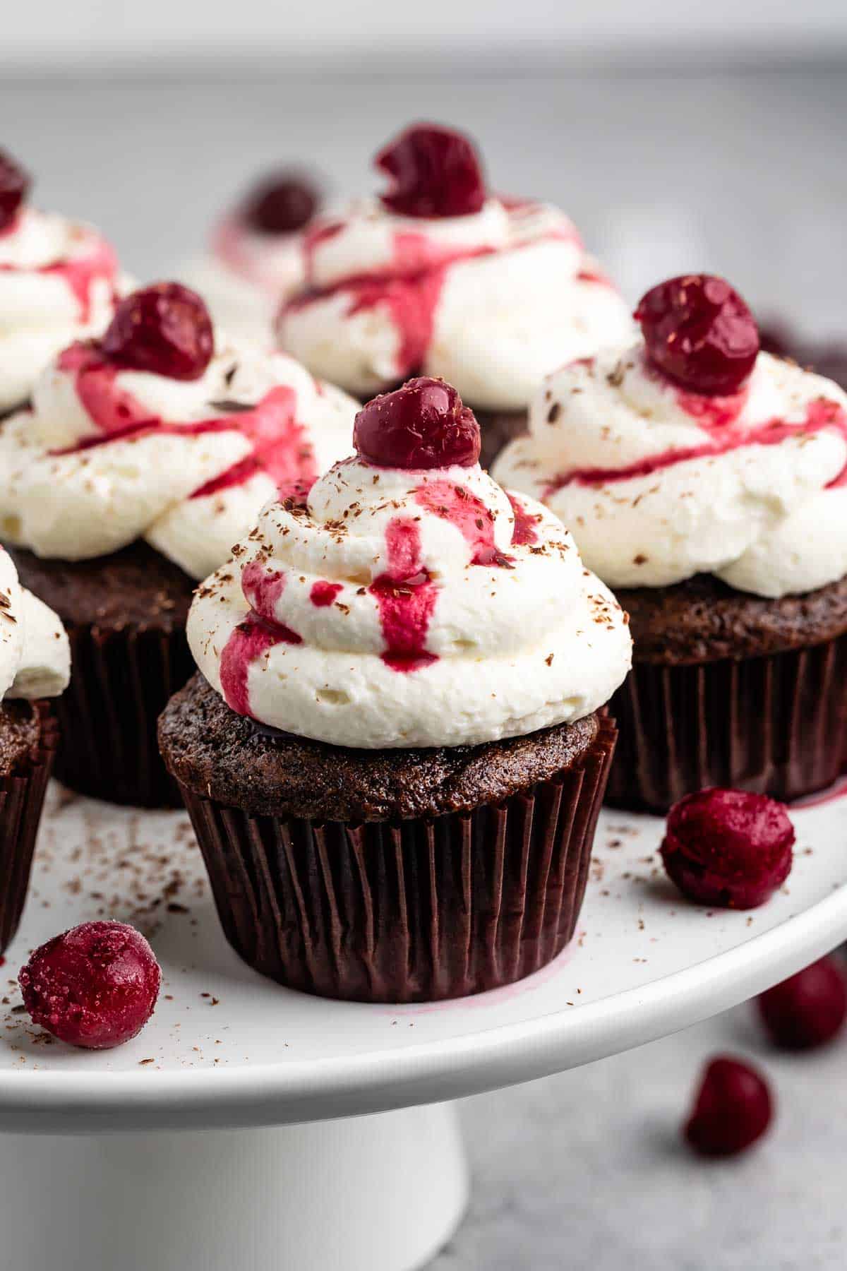 Chocolate cupcakes topped with swirls of white frosting, a red cherry on top, and drizzles of cherry sauce are arranged on a white cake stand. Small chocolate shavings and cherries are scattered around for decoration.