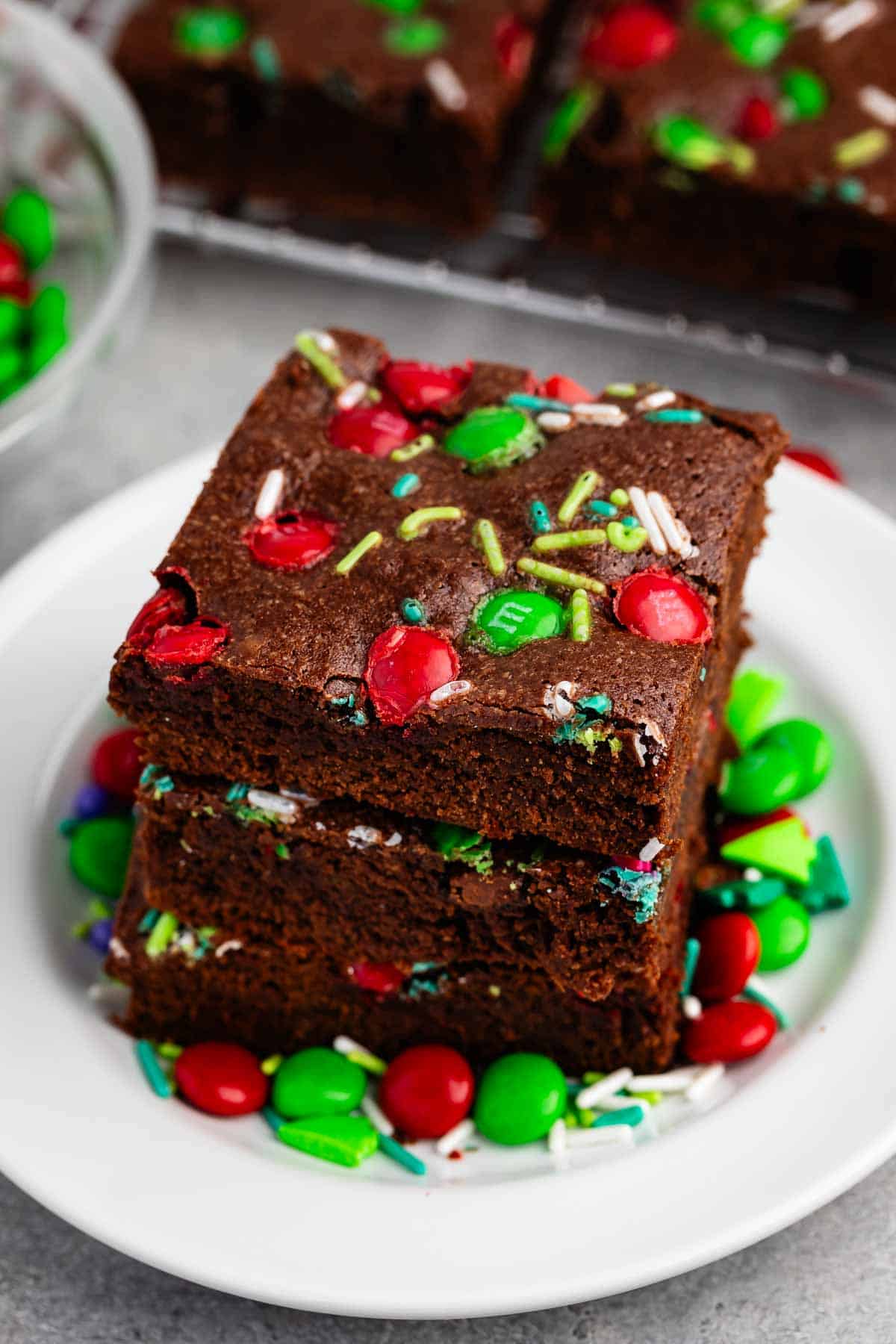 A stack of three chocolate brownies topped with colorful candy-coated chocolates and sprinkles sits on a white plate. More candies are scattered around the plate, with additional brownies visible in the background.