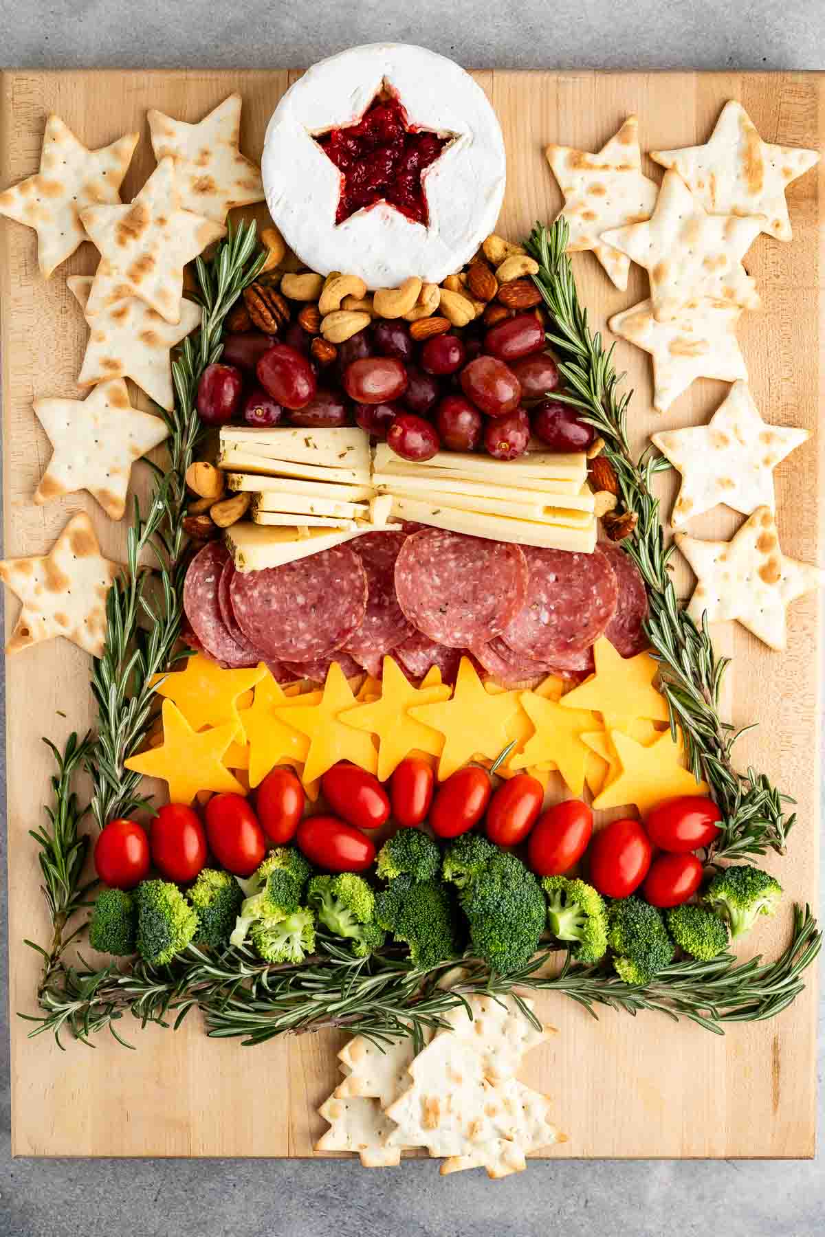 A festive charcuterie board arranged in the shape of a Christmas tree, featuring a brie with a star-shaped jelly, cheese slices, salami, grape tomatoes, broccoli, grapes, and rosemary sprigs. Surrounded by star-shaped crackers.