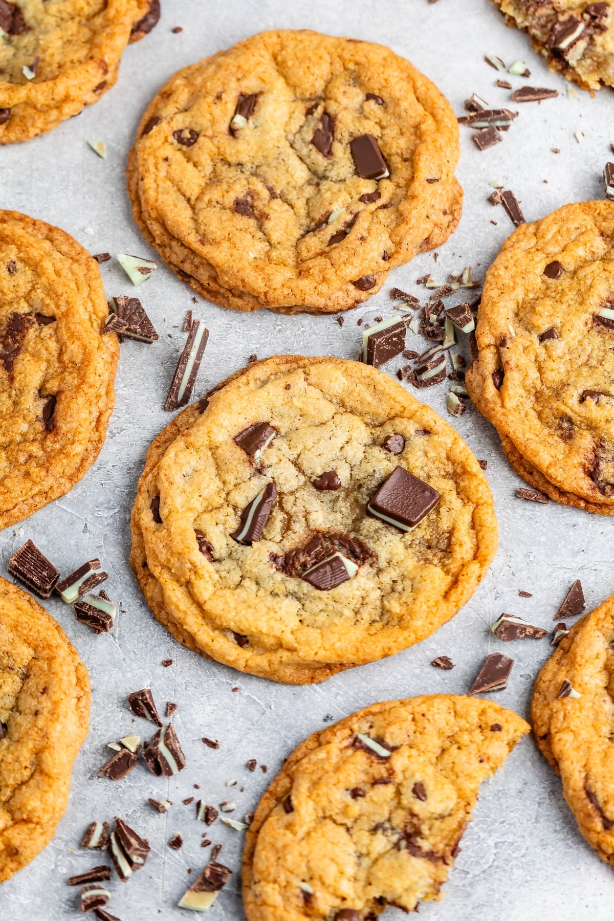 A group of freshly baked chocolate chip cookies with chunks of chocolate on top, arranged on a light gray surface. Some chocolate pieces are scattered around, enhancing the homemade and cozy appearance of the cookies.