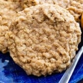 A close-up of a homemade peanut butter oatmeal cookie on a blue plate, with a spoonful of peanut butter beside it. More peanut butter oatmeal cookies are partially visible in the background.