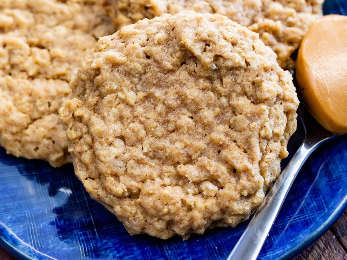 A close-up of a homemade peanut butter oatmeal cookie on a blue plate, with a spoonful of peanut butter beside it. More peanut butter oatmeal cookies are partially visible in the background.