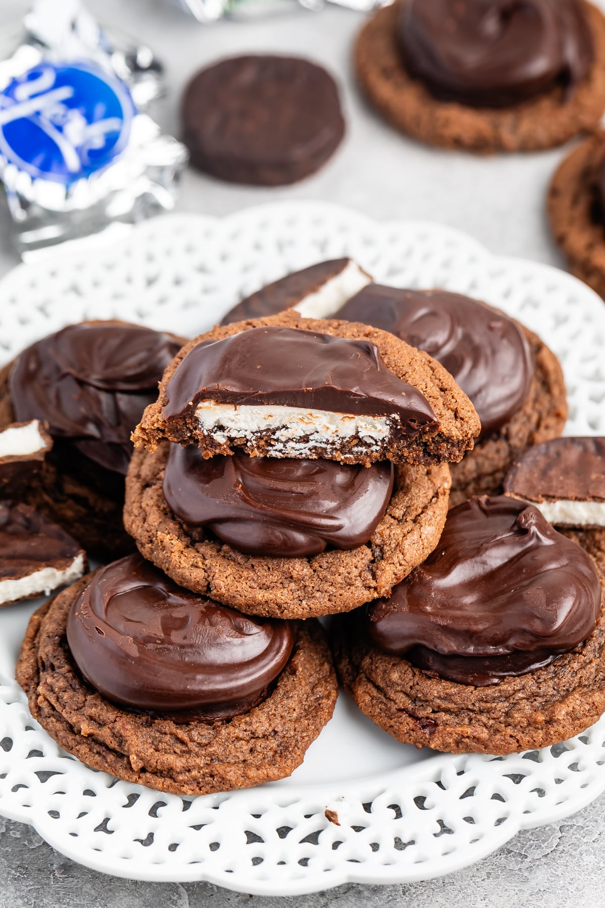 A plate of chocolate-covered cookies is stacked, with one cookie in the center broken in half to reveal a white filling. The cookies have a glossy chocolate coating and sit on a decorative white plate. Some wrapped cookies are blurred in the background.