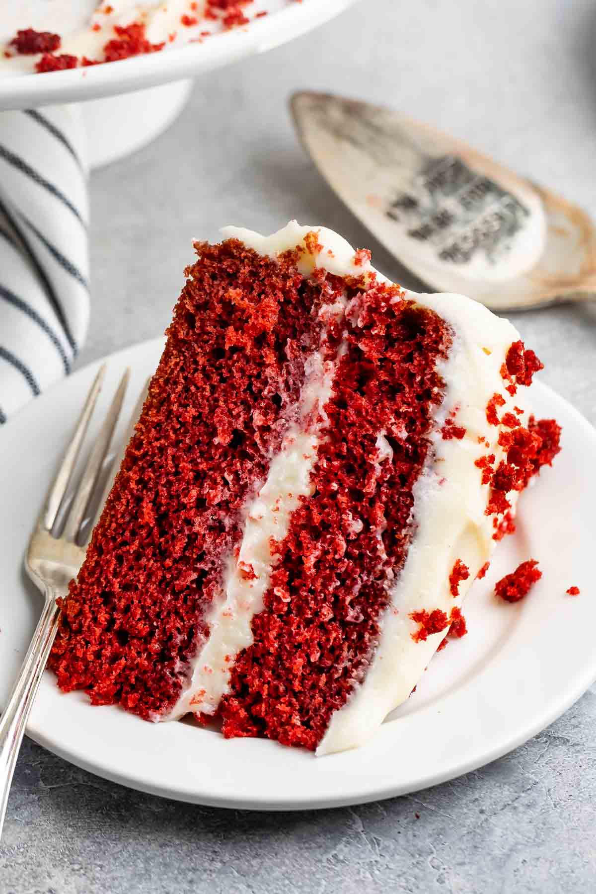 A slice of red velvet cake with creamy white frosting sits on a white plate. A fork is placed next to the cake, and a striped napkin is partially visible in the background.