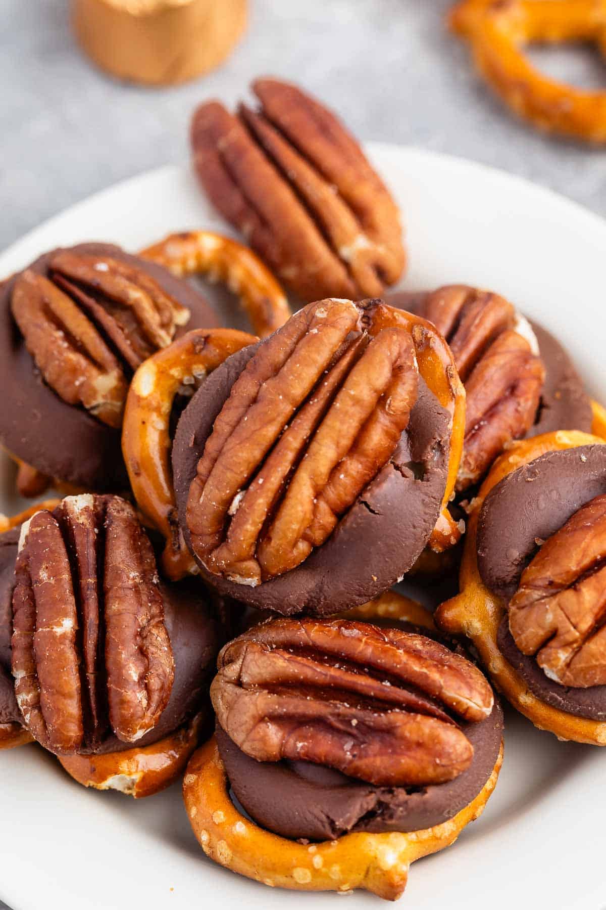 A plate of chocolate-covered pretzels topped with salted pecans. The treats are stacked on a white plate, showcasing a mix of smooth chocolate, crunchy pretzels, and whole pecans.