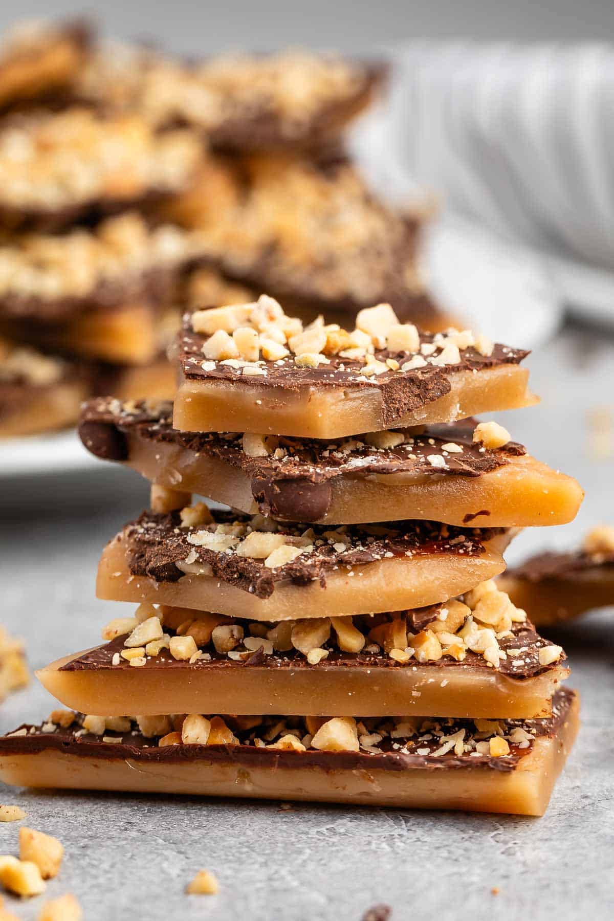 A stack of toffee pieces topped with chocolate and crushed nuts is placed on a gray surface. More toffee pieces are blurred in the background, giving a delicious and homemade dessert vibe.