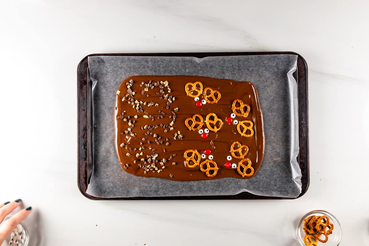 A baking tray with melted chocolate topped with pretzels, candy eyes, and chocolate chips is placed on parchment paper. A hand and a small bowl of pretzels are nearby on a white surface.