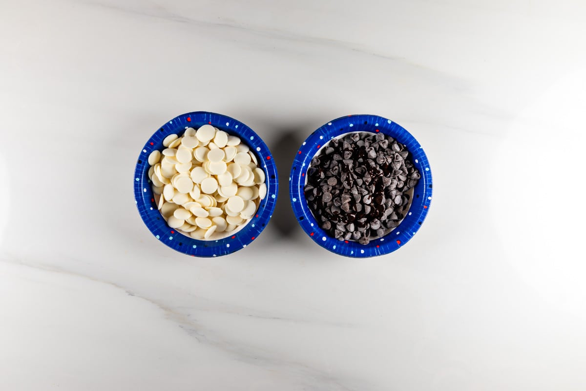 Two blue bowls on a light countertop, one filled with white chocolate chips and the other with dark chocolate chips.